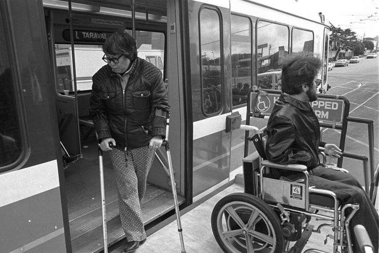 Man with crutches boarding bus while man in wheelchair waits on accessible platform, 1983.