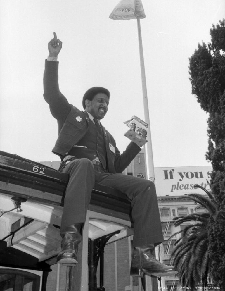 Man in uniform sitting atop cable car 62, raising fist in celebration while holding trophy.