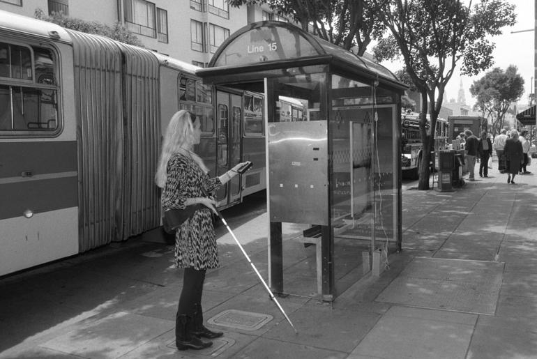 Woman with white cane using talking shelter sign at Line 15 bus stop with articulated bus nearby.