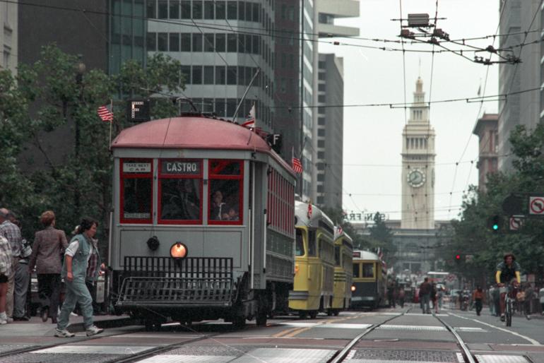 Red and yellow historic streetcars on Market Street with Ferry Building clock tower in background.