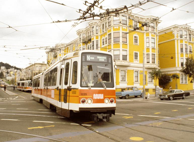 Muni light rail vehicle number 1225 at Church Street and Duboce Avenue intersection with yellow building in background.