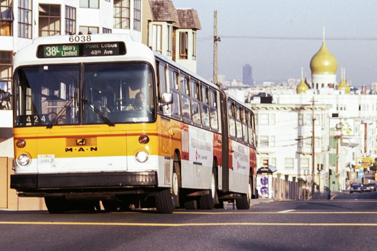 Yellow and white articulated bus on Geary Boulevard with church domes visible in background.