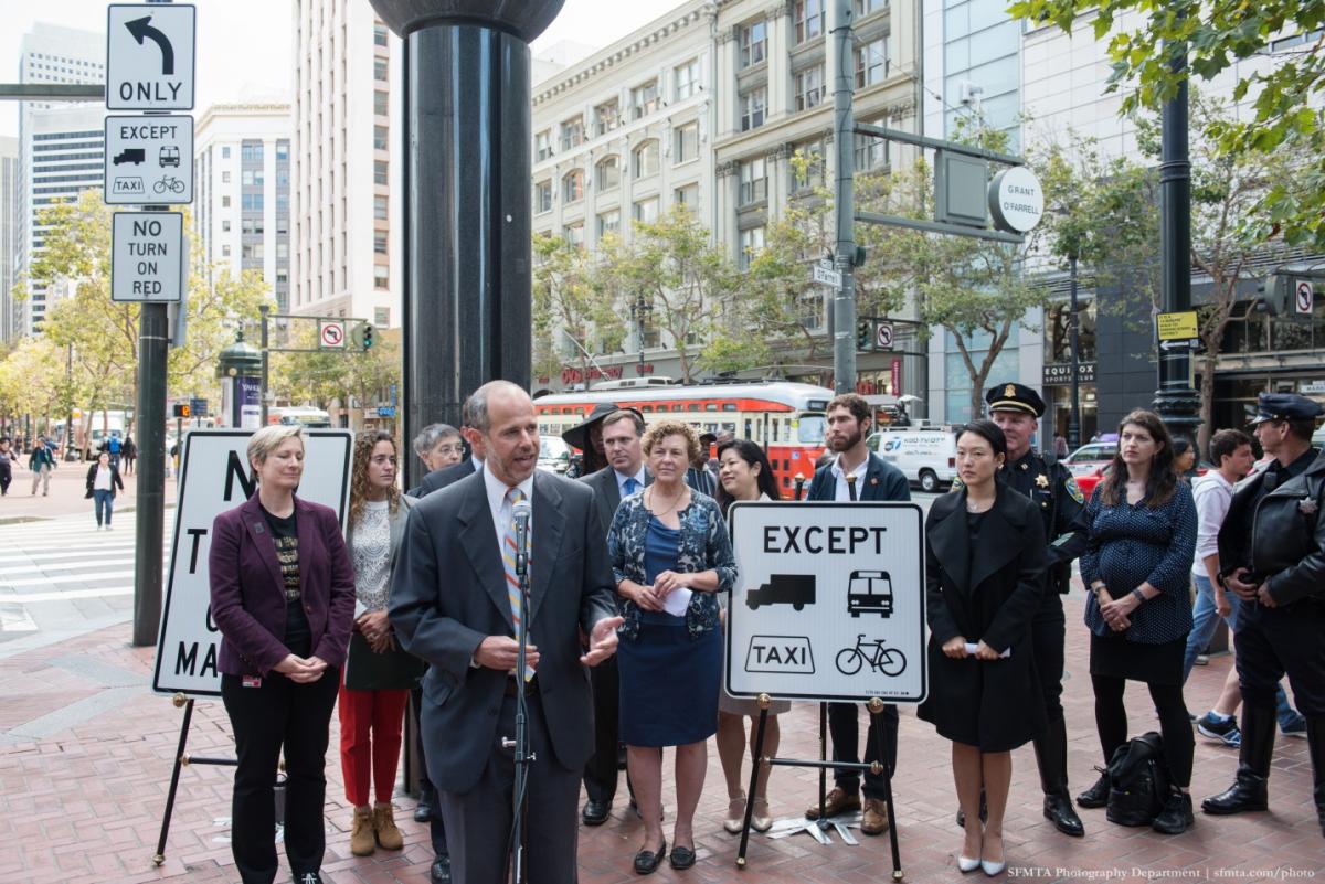 Man in suit speaking at microphone during downtown press conference with attendees holding traffic signs.