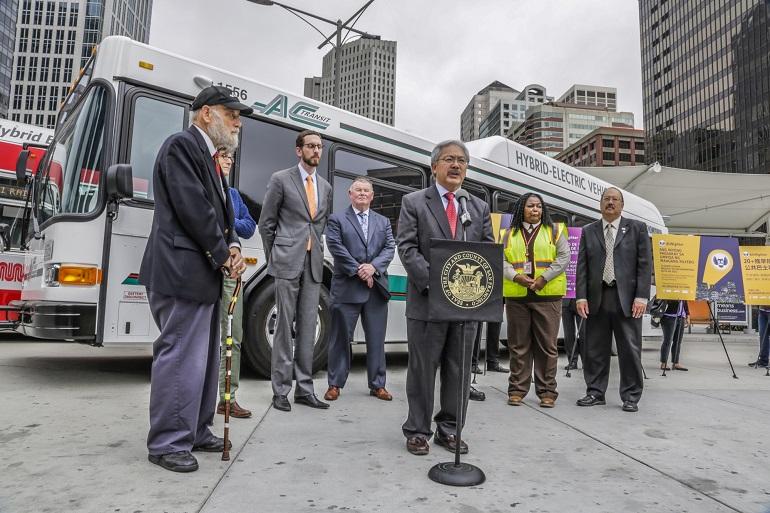 Officials and transit leaders stand at a press event in front of hybrid-electric buses at the Transbay Terminal.