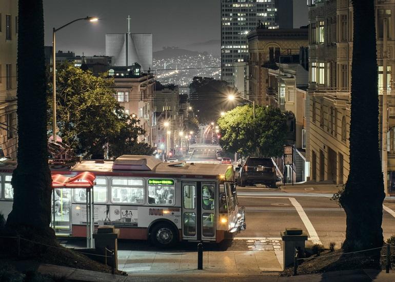 Muni bus stopped on a San Francisco street at night with city lights and buildings in background.