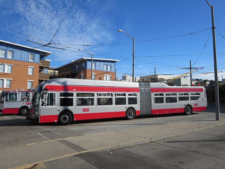 Red and white articulated trolley bus stopped at a Muni transit station with overhead electric lines visible.