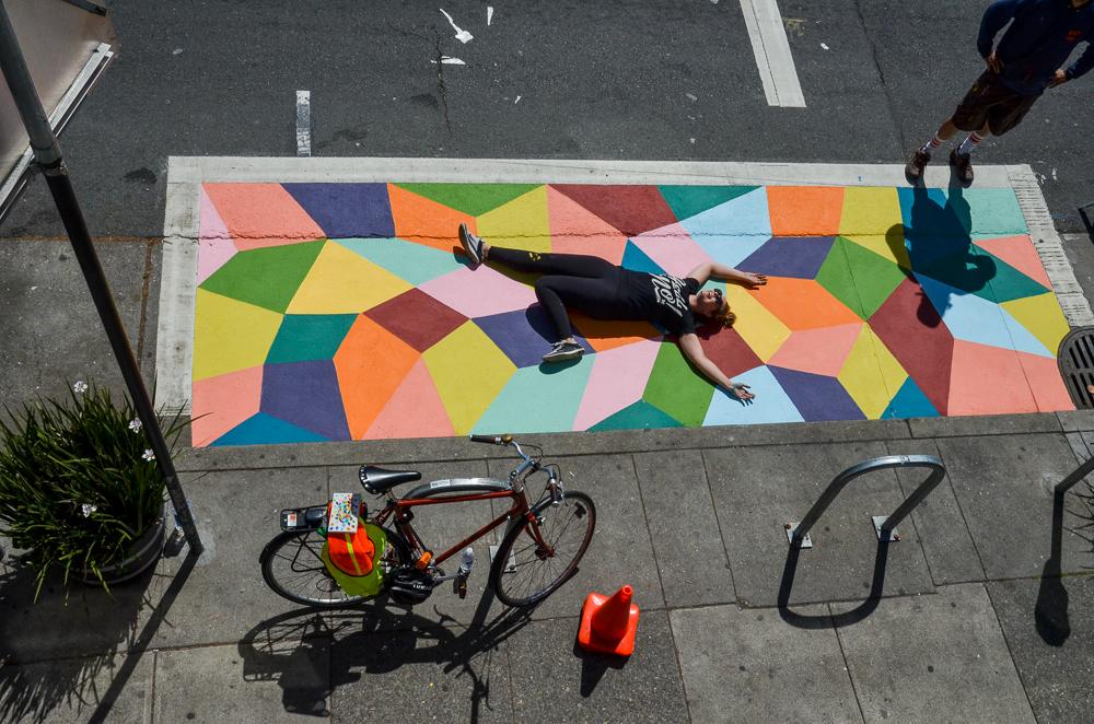 Person lying on colorful geometric mural next to bicycle parked on sidewalk.
