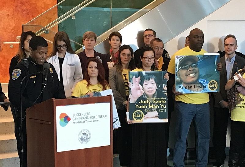 Police Chief William Scott at podium with city officials and advocates holding victim photos at Zuckerberg San Francisco General Hospital event.