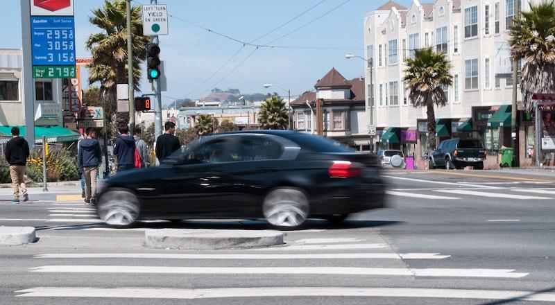 Black car in motion through crosswalk with pedestrians and San Francisco street scene in background.