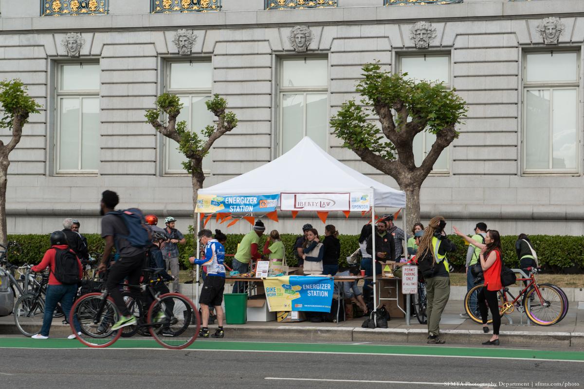Cyclists gather at a white pop-up tent energizer station on Polk Street in front of City Hall during Bike to Work Day.