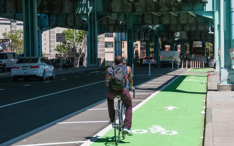 Cyclist riding on green protected bike lane under highway overpass in urban area.