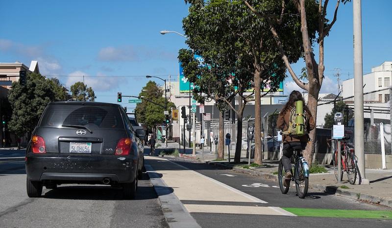 Cyclists ride on a protected bike lane next to parked cars on a city street with trees and buildings.