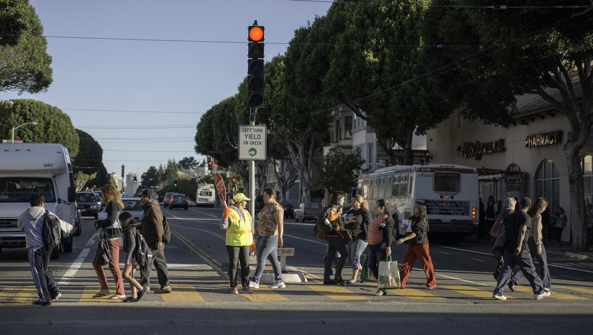 Crossing guard in yellow vest directs pedestrians and students across street at busy intersection with traffic light.