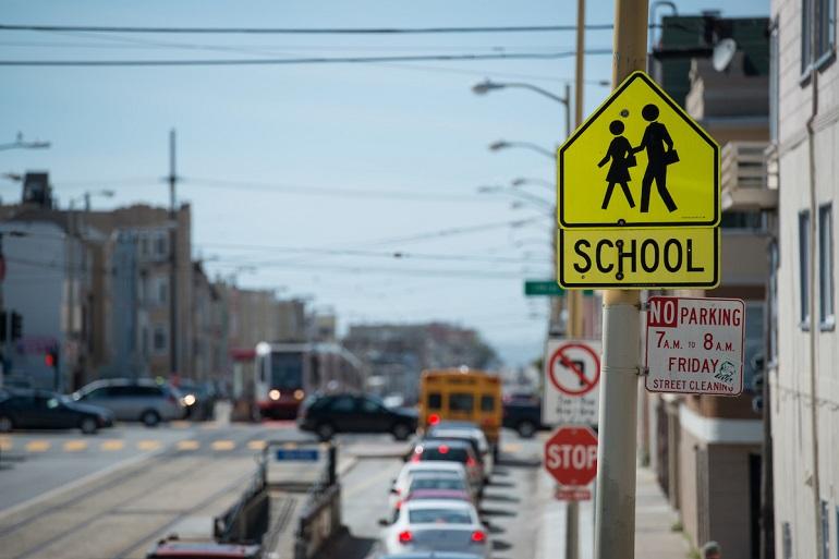 Yellow school crossing sign on urban street with parked cars and buildings.