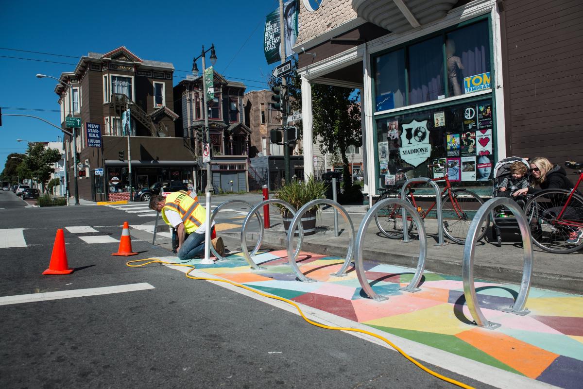 MTA worker installing circular bike rack above colorful geometric street mural with traffic cones nearby.