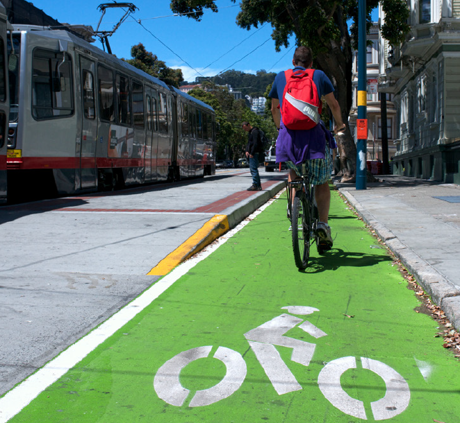 Cyclist riding on green protected bikeway next to cable car and yellow curb on city street.