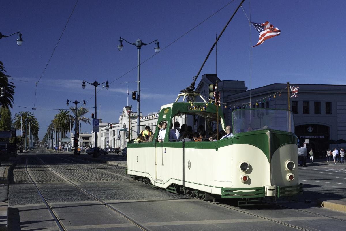 Green and white open-top streetcar traveling on tracks along The Embarcadero with American and British flags flying.