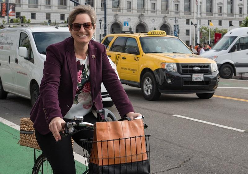 Woman on bicycle with orange bag in front of yellow taxi and City Hall building.
