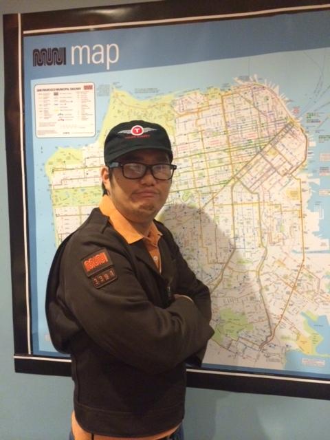 Muni operator in uniform standing in front of a colorful transit service map display.