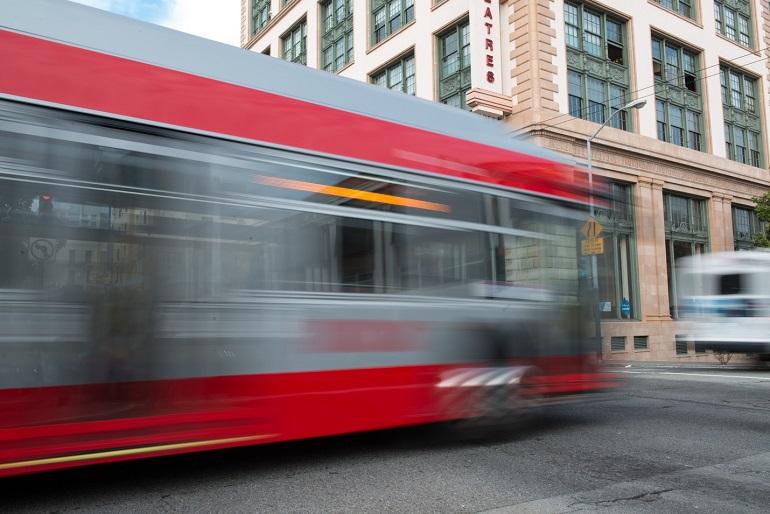Red and silver Muni bus in motion on downtown San Francisco street with historic building in background.