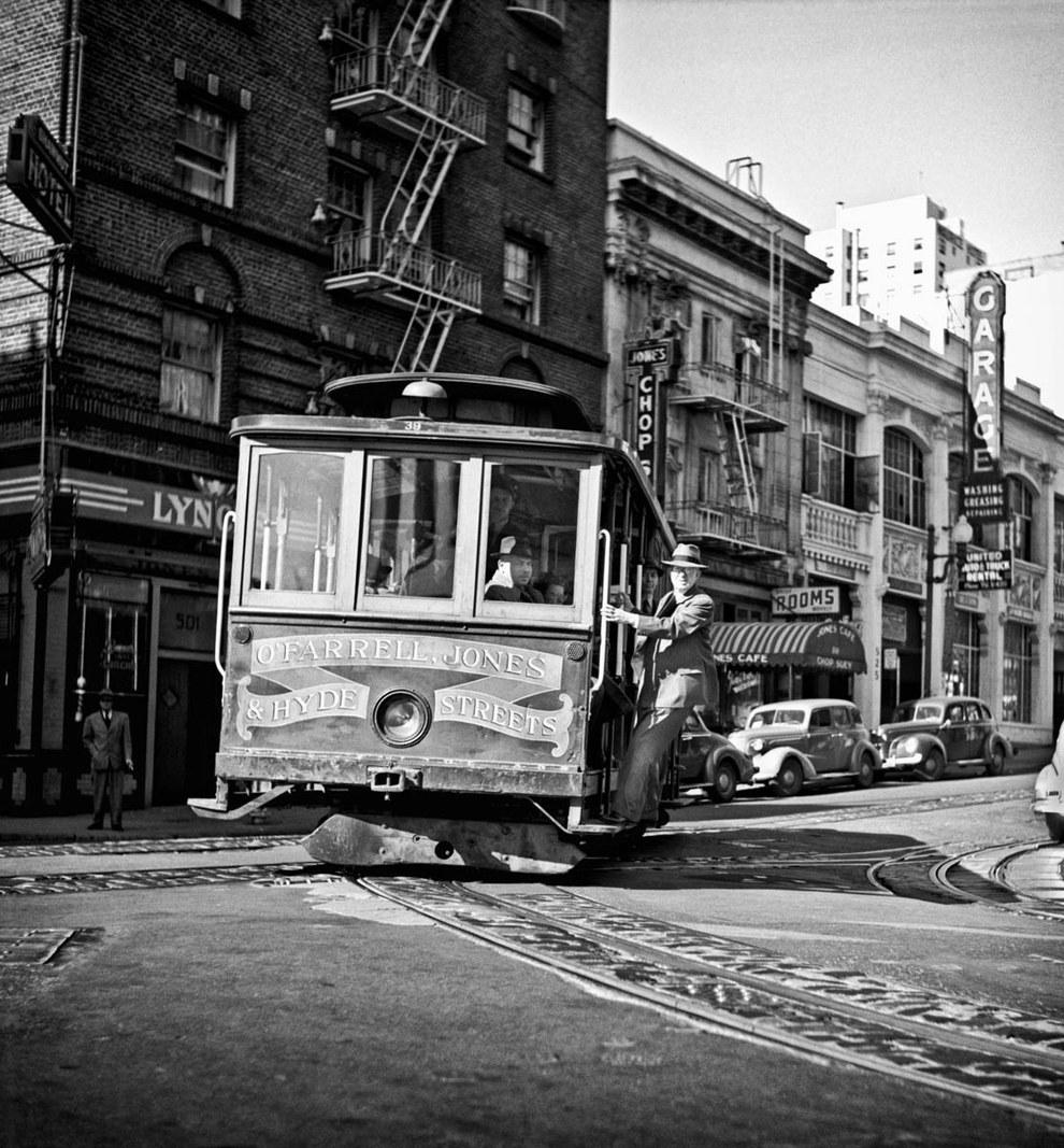 Cable car turning corner on San Francisco street with operator and passenger visible, vintage automobiles parked nearby.