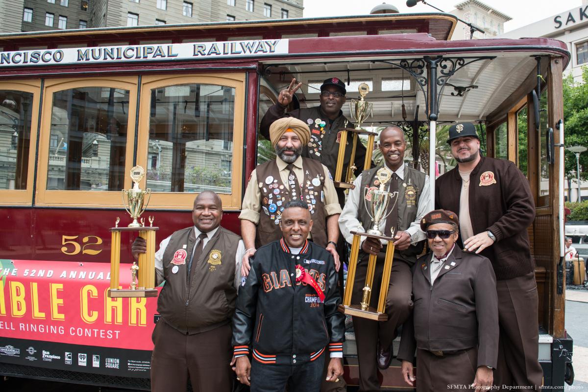 Seven men in brown Muni uniforms stand with trophies on a San Francisco cable car during Bell Ringing Contest celebration.