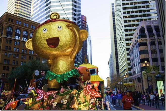 Large gold smiling monkey statue on parade float with green garland, surrounded by tall buildings on Market Street.