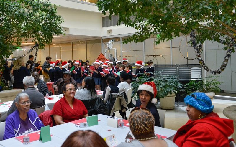 People seated at tables during a holiday celebration with a children's choir performing in the background.
