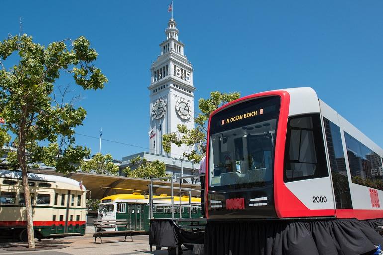 Red and white light rail vehicle displayed at The Embarcadero with Ferry Building clock tower in background.