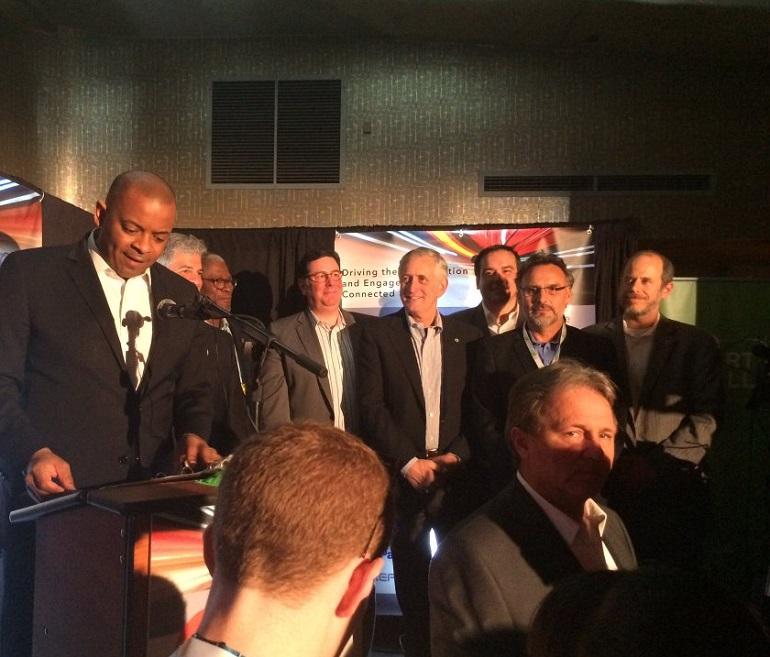 Man speaking at podium with seven city representatives standing behind him at conference event.