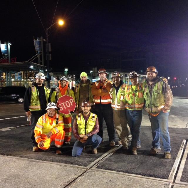 Construction crew members in safety vests and hard hats pose at night near Caltrain station during track installation work.