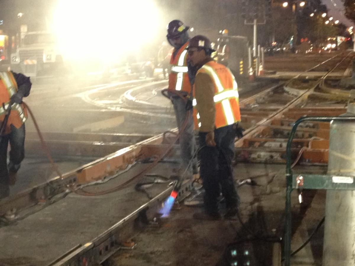 Crew members in safety vests and hard hats work on track installation at night, with welding equipment visible.