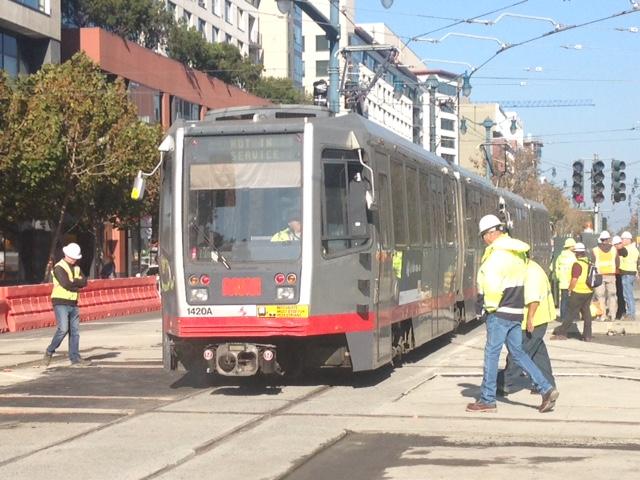 Construction workers in safety vests stand beside a Muni light rail train during track testing on 4th Street.