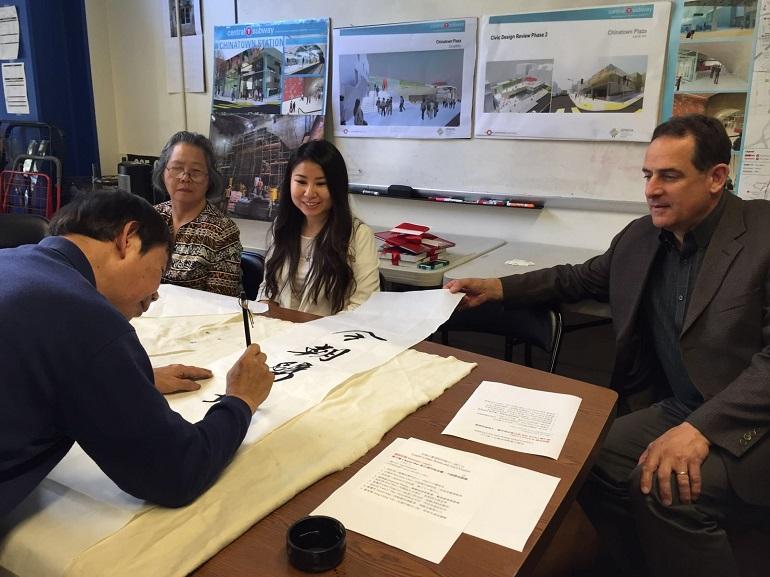 Calligraphy artist writes characters on paper while four people observe during Central Subway couplet press event.
