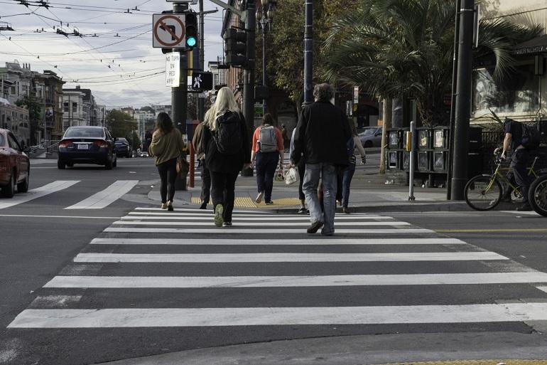 Pedestrians crossing striped crosswalk on busy urban street with vehicles and palm trees visible.