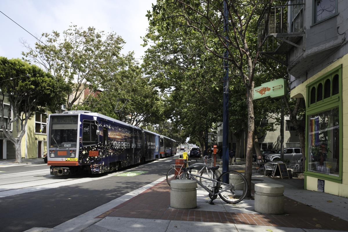 Light rail vehicle with dark blue wrap traveling on tree-lined street past storefronts and parked bicycle.