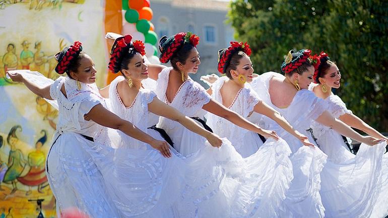 Six female dancers in white dresses with red flowers in their hair performing a traditional Mexican folk dance outdoors.