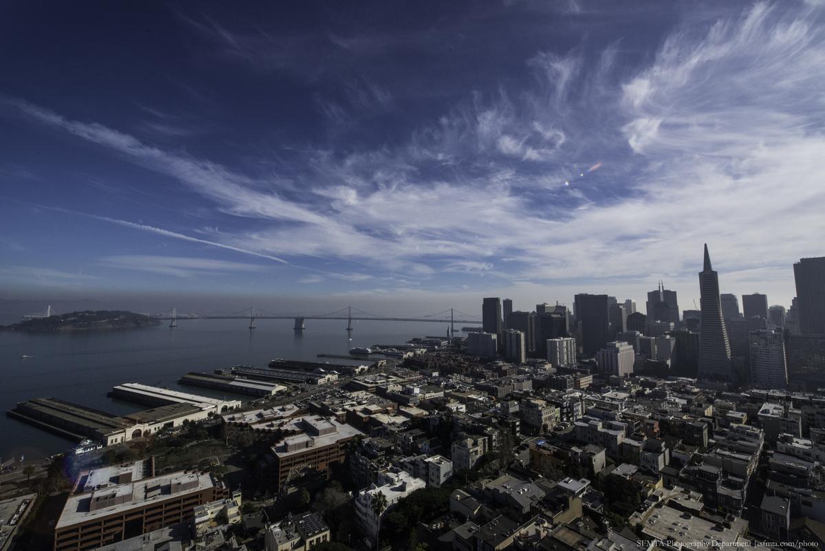 Aerial view of San Francisco skyline with bay, bridges, and waterfront piers under blue sky with wispy clouds.