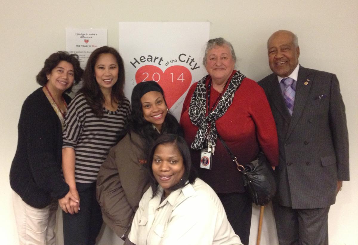 Six people stand in front of a 2014 Heart of the City Combined Charities Campaign poster.