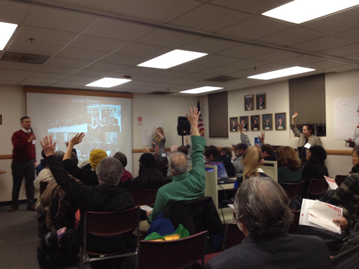 Seated audience members raise hands during a community meeting in a classroom with a projection screen.