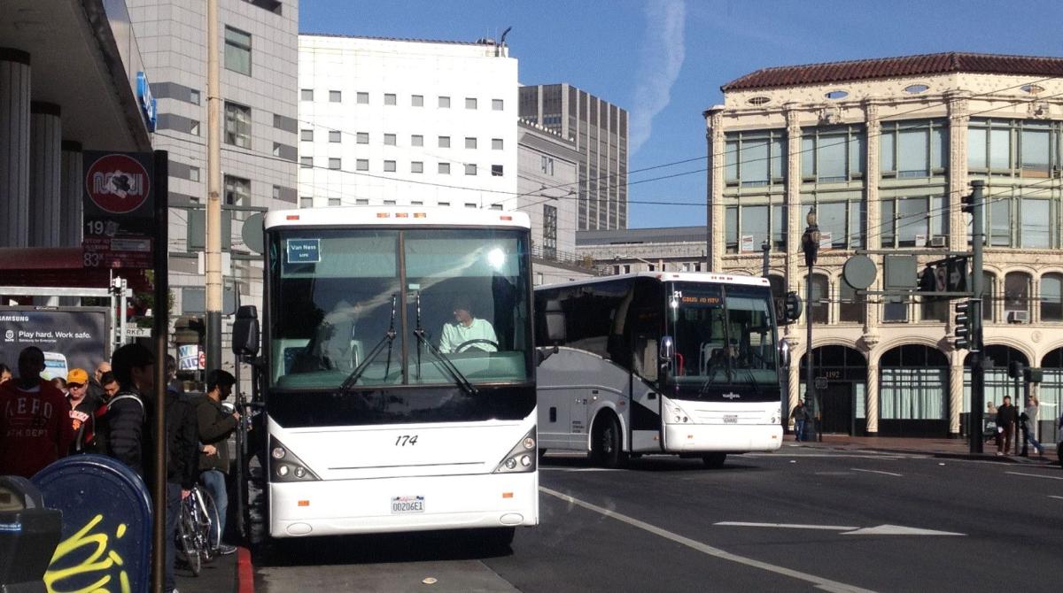 Two white buses stopped at a busy urban street corner with pedestrians and historic buildings.