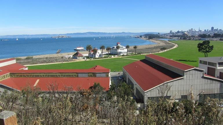 Crissy Field with red-roofed buildings, green lawn, and San Francisco Bay with city skyline in background.