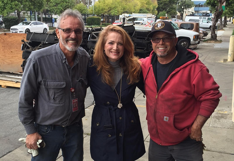 Three people smile at camera on sidewalk with construction vehicles and parked cars in background.
