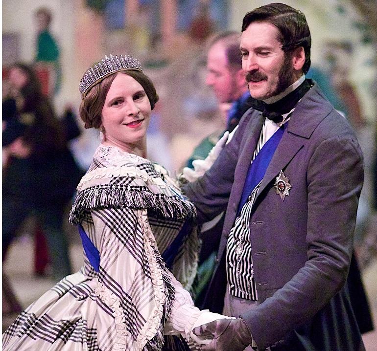 A couple in Victorian-era formal attire dancing together at an indoor event with spectators in the background.
