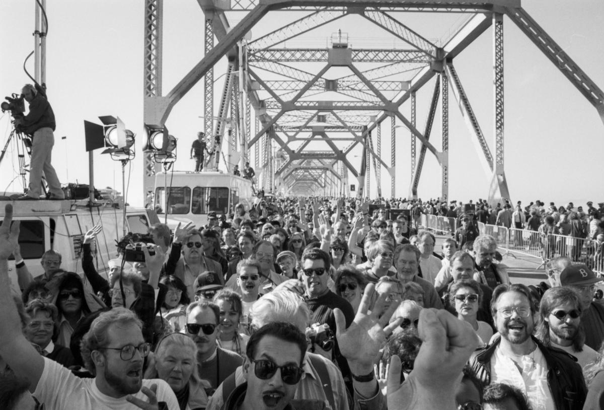 Crowds of people walking across the Bay Bridge's upper deck with TV cameras and lighting equipment visible on the left.