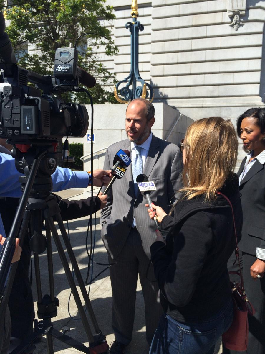 Man in gray suit speaks to reporters with microphones in front of City Hall building.