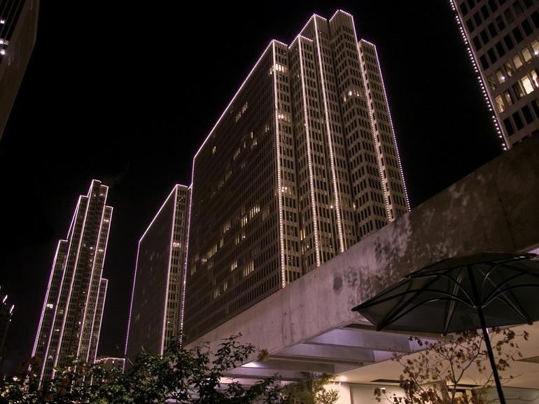 Illuminated skyscrapers at night with pink-outlined architectural lighting against dark sky, modern building facade below.