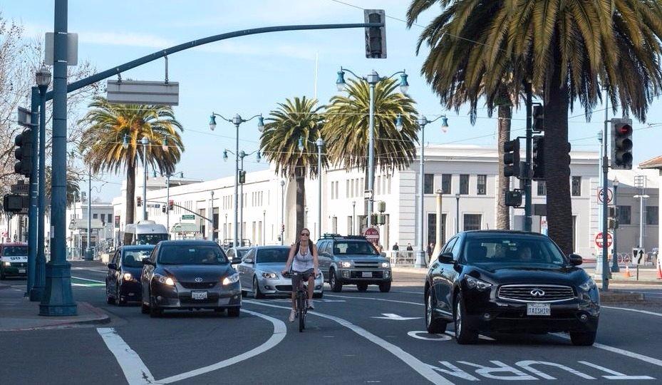 Cyclist riding in bike lane on Embarcadero with palm trees, traffic, and historic buildings visible.