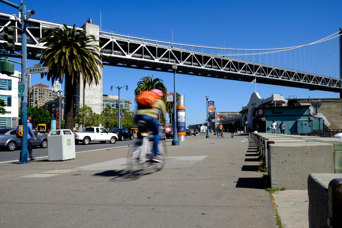 Bicyclist riding along The Embarcadero with Bay Bridge and waterfront buildings visible under clear blue sky.