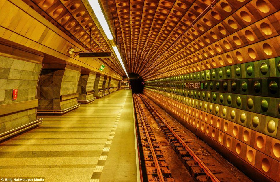 Malostranská metro station platform with golden-yellow tiled walls and circular perforations creating a sunburst perspective down the tunnel.